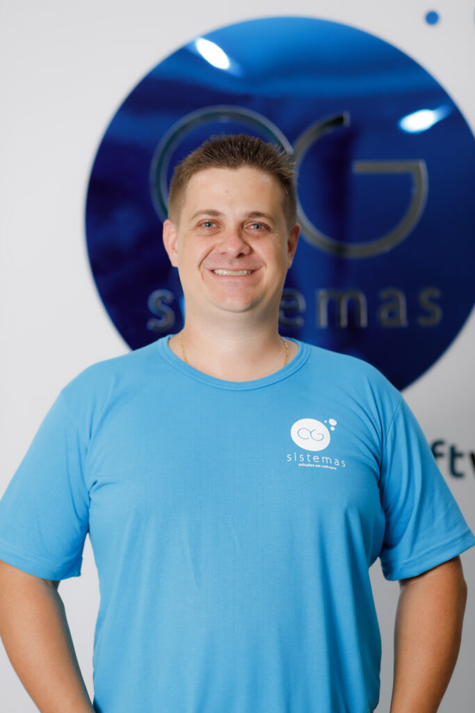 Rafael Rotoli, um dedicado desenvolvedor da AG Sistemas, está sorridente e confiante em frente ao logo da empresa. Ele veste uma camisa azul com o logotipo da AG, simbolizando seu pertencimento e paixão pelo trabalho que contribui para seu crescimento pessoal e profissional.