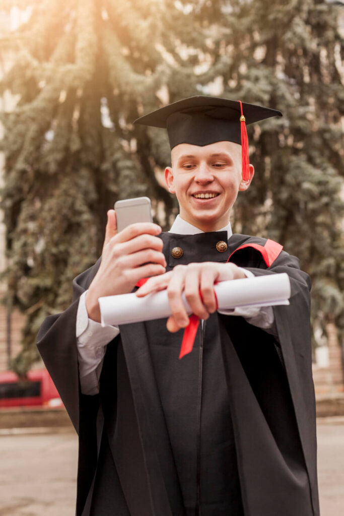 Formando em traje acadêmico segura um diploma amarrado com uma fita vermelha em uma mão e um smartphone na outra, ao ar livre com árvores e luz solar ao fundo, rosto pixelado para privacidade.”