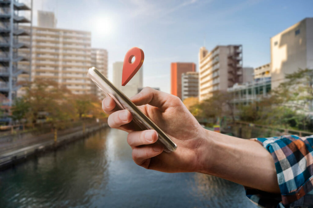 Mão segurando um smartphone com ícone de localização, em um cenário urbano, representando as Estratégias Imobiliárias Inovadoras no contexto da tecnologia e navegação imobiliária.