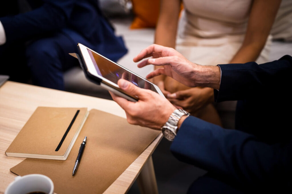 Duas pessoas sentadas em uma mesa de reunião, com uma delas interagindo com um tablet, ao lado de um caderno e uma caneta, enfatizando ‘A Importância do Cadastro Correto de Produtos’ em um contexto profissional.