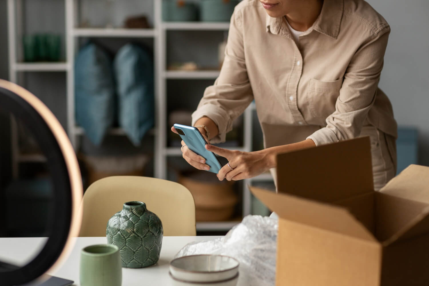 Uma pessoa concentrada no cadastro de produtos com um smartphone, ao lado de uma caixa com itens de cerâmica, ressaltando ‘A Importância do Cadastro Correto de Produtos’ para o controle de inventário e vendas online.