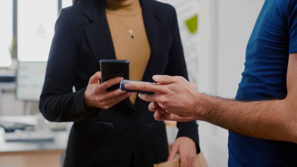 Duas pessoas em um ambiente de escritório, com foco nas mãos. Uma pessoa, vestindo uma blusa preta e casaco, segura um smartphone. A outra pessoa, com uma camisa azul e mangas curtas, aponta para a tela do smartphone. A interação sugere uma troca de informações ou demonstração de produto que pode estar relacionada à frase-chave ‘Revolucione Suas Vendas de Álbuns’, indicando inovação ou modernização no processo de vendas.