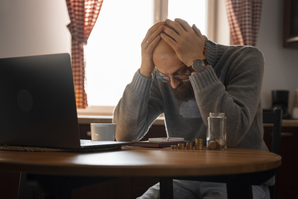 Homem com as mãos na cabeça, demonstrando preocupação ao olhar para a mesa com moedas e um laptop, refletindo os desafios financeiros. A inadimplência está sufocando o seu negócio?