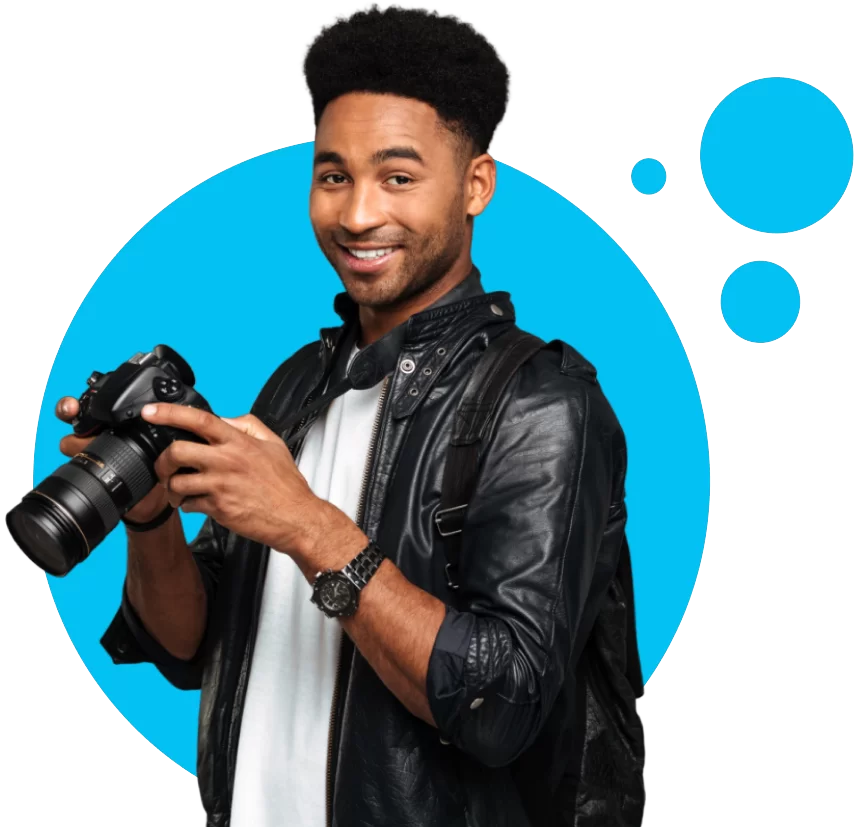 Fotógrafo sorridente com camiseta azul e jaqueta de couro, segurando uma câmera DSLR, pronto para capturar momentos memoráveis numa festa de formatura personalizada. Personalização de Festas de Formatura.
