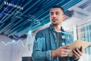 A imagem ilustra um homem jovem e contemplativo, vestindo uma camisa casual azul, engajado na análise de estatísticas financeiras proeminentes que flutuam à frente dele como se numa interface futurística de realidade aumentada. Ele segura um tablet, que parece ser o dispositivo através do qual interage com os dados virtuais. Os elementos gráficos destacam importantes métricas de negócios, incluindo saldo total, rendimentos e despesas, sublinhando a habilidade do sistema ERP da AG Sistemas em fornecer análises detalhadas e em tempo real para informar a tomada de decisões estratégicas. A atmosfera da imagem é moderna e tecnologicamente avançada, alinhando-se perfeitamente com a temática da capa sobre "Tendências futuras em sistemas ERP".