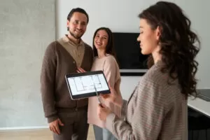 Um jovem casal sorridente observa uma planta de imóvel em um tablet, mostrado por uma agente imobiliária, destacando a praticidade e eficiência da tecnologia digital no processo de visualização de propriedades. A interação ocorre em um ambiente moderno e bem iluminado, simbolizando uma abordagem inovadora na apresentação de imóveis.