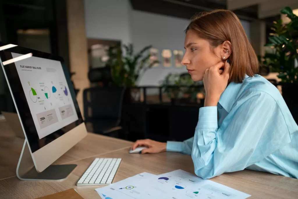 Uma pessoa está sentada em uma mesa de escritório, olhando para um monitor de computador que exibe gráficos coloridos e análises financeiras. Documentos estão espalhados na mesa, e a cena transmite a ideia de “Simplificando gestão financeira” através da organização visual e apresentação clara dos dados financeiros, facilitando a tomada de decisões e o gerenciamento de informações complexas.