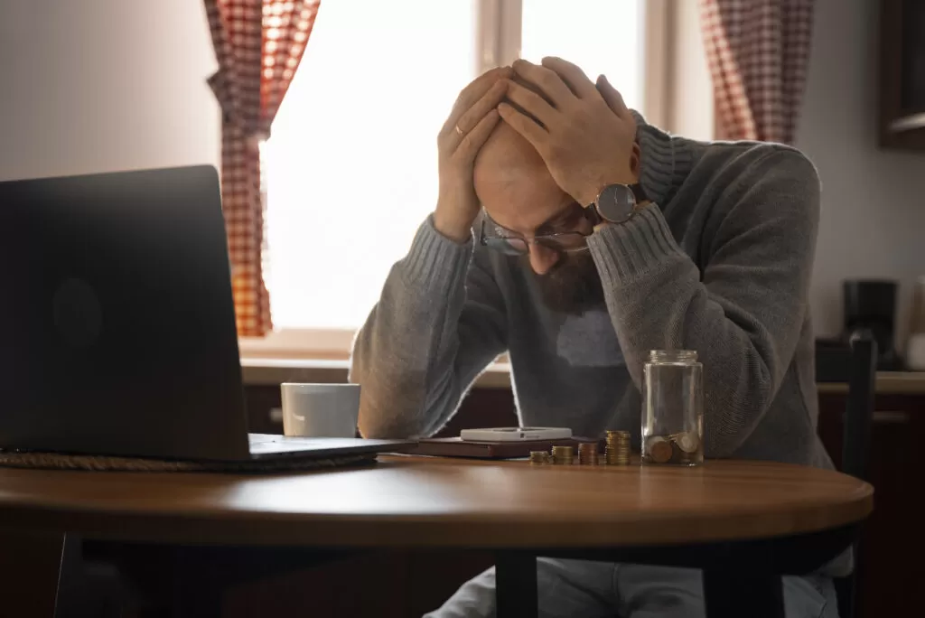 Homem com as mãos na cabeça, demonstrando preocupação ao olhar para a mesa com moedas e um laptop, refletindo os desafios financeiros. A inadimplência está sufocando o seu negócio?