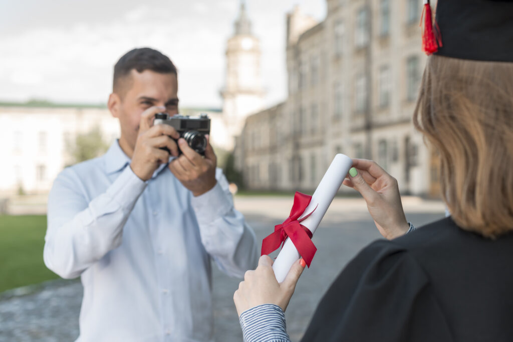 A imagem mostra uma pessoa vestida com trajes de formatura segurando um diploma com uma fita vermelha, sendo fotografada por outra pessoa com o rosto oculto. Isso simboliza um evento significativo que requer planejamento e organização cuidadosos para capturar e celebrar conquistas educacionais, destacando a importância da organização de agenda de eventos.