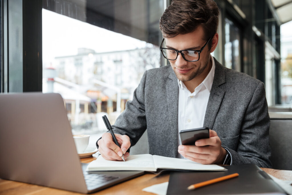 Empresário sorridente usando óculos, trabalhando em um café com laptop e smartphone, gerenciando tarefas e escrevendo notas, simbolizando a eficiência na gestão de boletos e organização financeira.