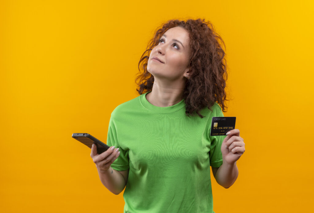 Mulher segurando um celular e um cartão de crédito, olhando pensativa, ilustrando 5 estratégias para evitar inadimplência.