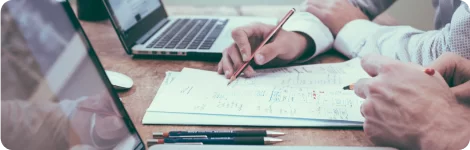 Duas pessoas colaborando em uma mesa de escritório, com foco em suas mãos escrevendo e revisando notas em um papel com gráficos e listas. Um laptop aberto, canetas e outros materiais de escritório são visíveis, indicando um ambiente de trabalho produtivo. A imagem sugere uma reunião de planejamento ou uma sessão de estratégia financeira, alinhada com as funcionalidades do sistema agPhoto que apoia empresas de formaturas e eventos na gestão de contratos, vendas e análises financeiras, destacando a importância de um planejamento detalhado para o sucesso de eventos.