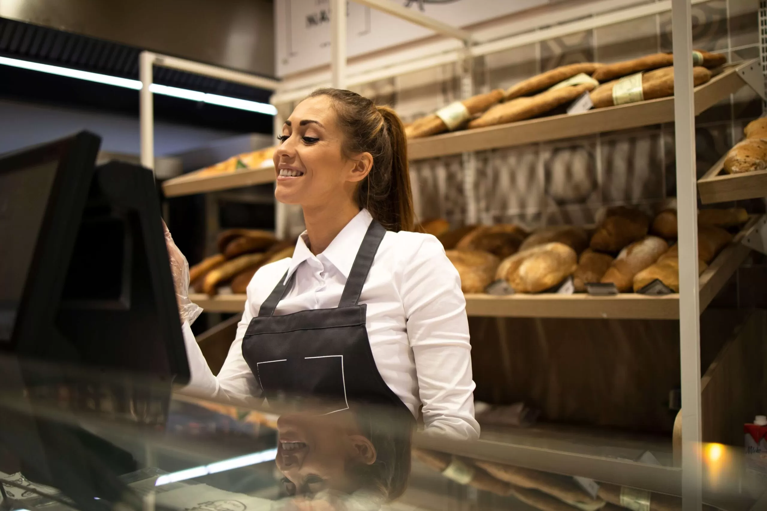 Uma vendedora sorridente em uma padaria, vestindo uma camisa branca e avental preto, interage com um sistema de computador provavelmente ligado a um ERP de gestão alimentar, com prateleiras ao fundo exibindo uma variedade de pães frescos iluminados por luzes LED. A imagem reflete a combinação de atendimento ao cliente amigável e tecnologia eficiente no setor de panificação.