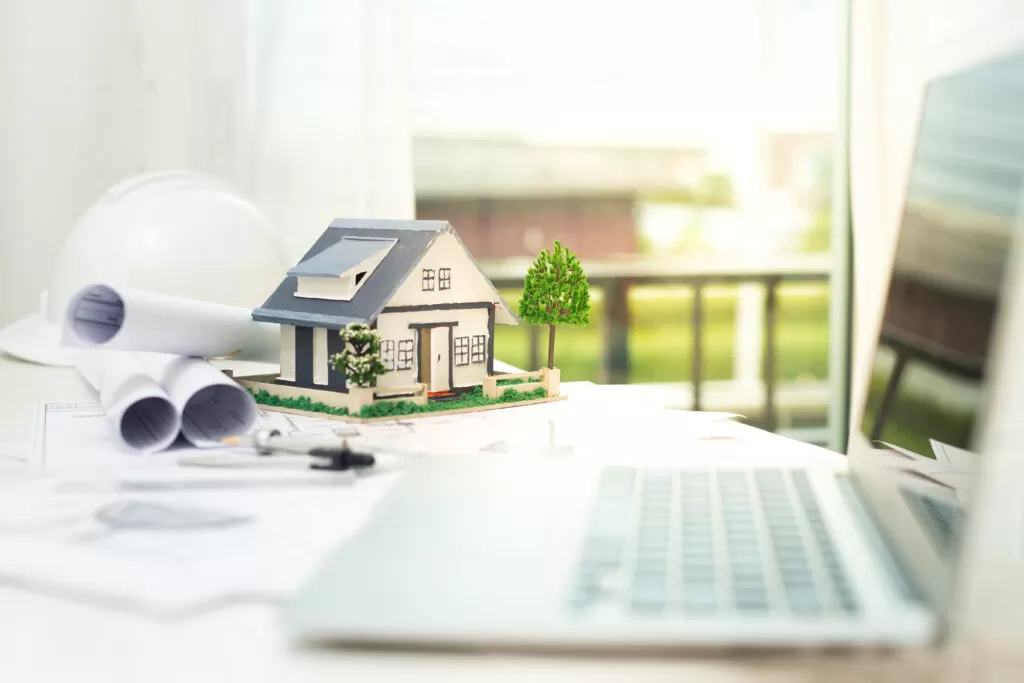 Obrigações das Imobiliárias na Lei do Inquilinato: Miniatura de casa em mesa de escritório, cercada por projetos e notebook, simbolizando a importância da conformidade legal na administração de imóveis.